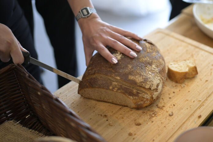 Auf dem Bild ist eine Person zu sehen, die ein rundes, dunkles Brot mit einer Kruste auf einem Holzbrett schneidet. Die linke Hand hält das Brot fest. In der rechten Hand hält die Person ein Brotmesser, um eine Scheibe abzutrennen.