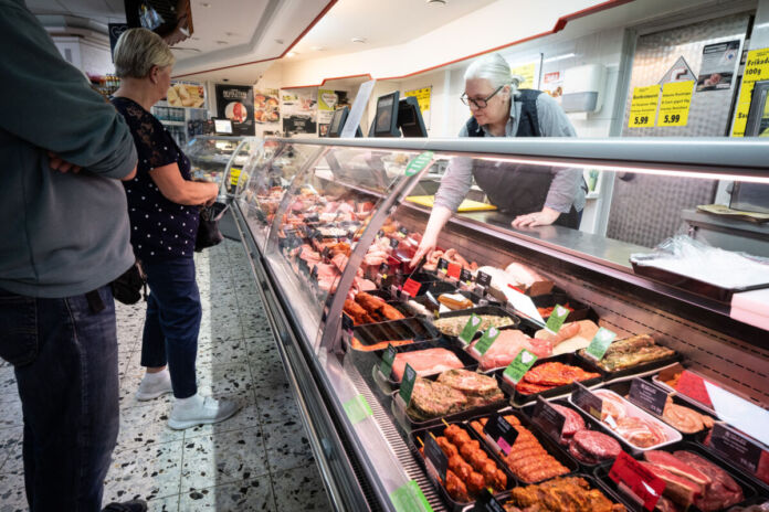 Vor einer Fleisch- und Wurstheke stehen mehrere Kunden und sehen sich die ausgelegten Fleisch- und Wurstwaren an. Eine Verkäuferin beugt sich gerade in die Theke und zeigt auf ein Stück Fleisch.