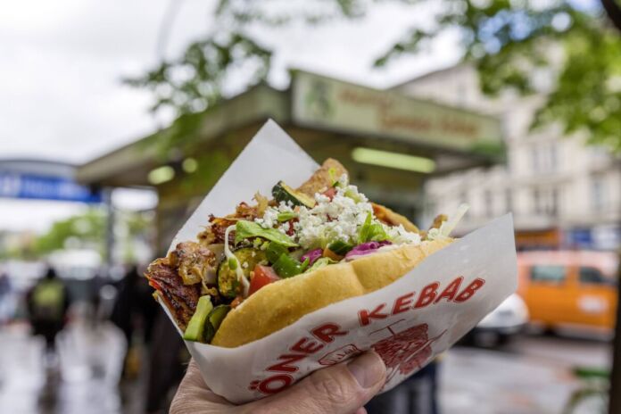 Eine Person hält einen Döner mit Fleisch, Gemüse und Fetakäse in der Hand. Im Hintergrund sieht man einen Imbiss an einer U-Bahn-Station.
