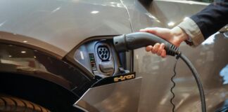 Ein Mann tankt und lädt gerade ein E-Auto mithilfe einer elektrischen Zapfsäule, einer Ladestelle auf. Er hält den Ladeschlauch in das Auto, damit die Batterie für mehr Reichweite voll aufgeladen wird.