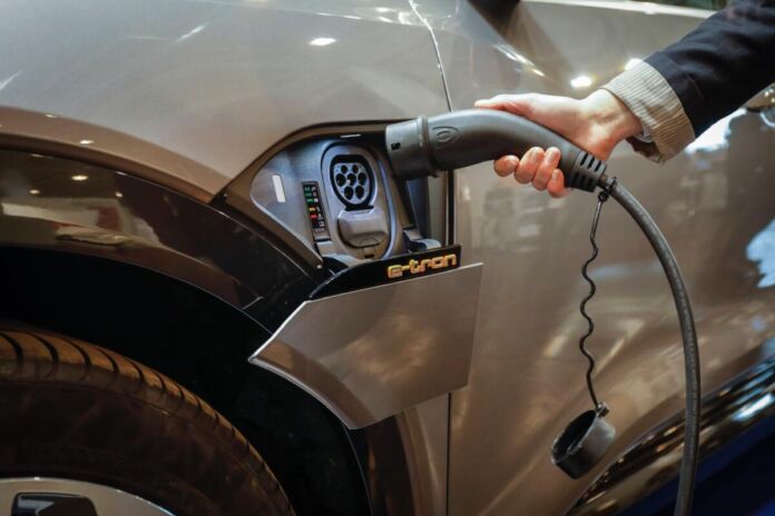 Ein Mann tankt und lädt gerade ein E-Auto mithilfe einer elektrischen Zapfsäule, einer Ladestelle auf. Er hält den Ladeschlauch in das Auto, damit die Batterie für mehr Reichweite voll aufgeladen wird.