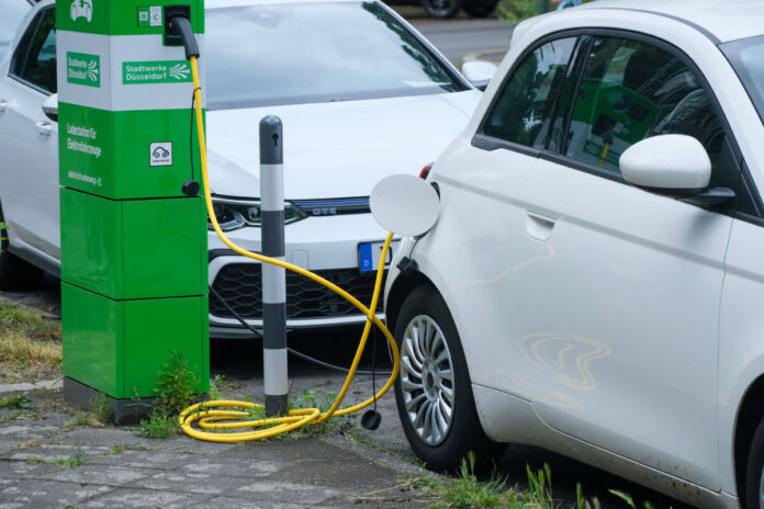 Ein weißes Elektroauto lädt gerade per Kabel, stehend, bei einer Elektrotankstelle an einer Strom-Zapfsäule auf, um wieder weiterfahren zu können. Dahinter parkt ein anderes weißes Fahrzeug.