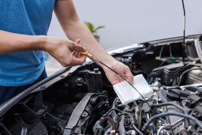 Ein Mechaniker im blauen Shirt führt eine Kontrolle am Motor eines Autos, eines Verbrenners durch. Er macht wohl mit seinem Gerät eine Öl-Probe und schaut, ob mit dem Fahrzeug alles stimmt.