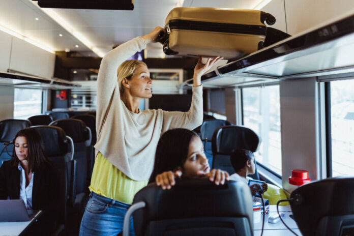 Eine Frau steht im Gang eines Zugabteils und hebt gerade ihren Koffer über den Köpfen der anderen Fahrgäste hinweg auf eine Ablagefläche. Eine Frau, die direkt davor sitzt, hält sich am Vordersitz fest und schaut gerade aus dem Fenster. Eine andere Frau arbeitet an einem Laptop.