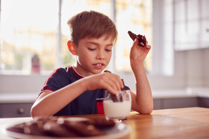 Ein Junge sitzt an einem Holztisch. In der linken Hand hält er einen angebissenen Keks. Mit der rechten tunkt er gerade einen weiteren Keks in ein Glas Milch ein. Im Vordergrund erkennt man verschwommen einen Teller mit weiteren Keksen.