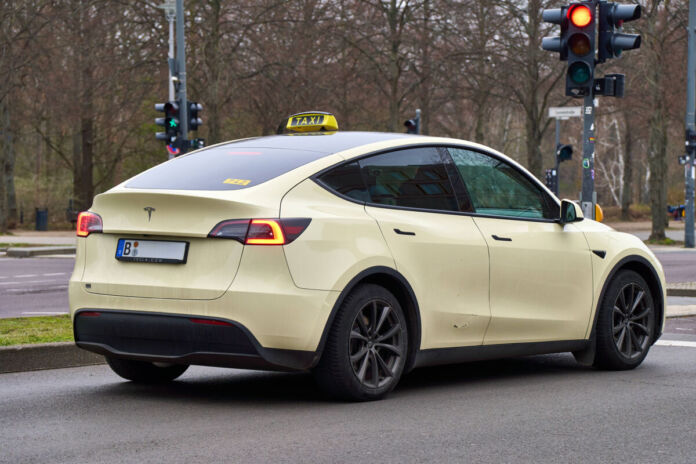 Auf einer Straße steht ein Taxi an einer roten Ampel. Es handelt sich um ein Tesla-Auto mit einem Berliner Kennzeichen. Die Bäume im Hintergrund sind kahl.