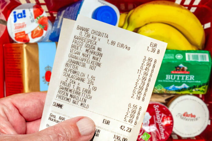 Die Hand eines Mannes hält einen Kassenzettel in der Hand. Darauf stehen die Produkte, die er offenbar gekauft hat, sowie die Preise. Im Hintergrund erkennt man einen vollen Einkaufskorb mit Lebensmitteln.