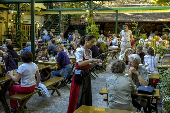 Sehr viele Gäste sitzen in einer Gaststube - sie werden auf der Terrasse von den Kellnern bedient und erhalten Speisen und Getränke. Die Stimmung ist traditionell und urig.