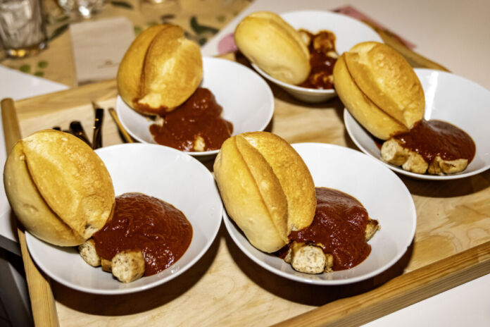 Auf einem Holztablett befinden sich offenbar fünf Kinderteller. Die Portionen sind klein. Auf jedem Teller befindet sich ein Brötchen mit einer geschnittenen Currywurst und entsprechender Sauce.