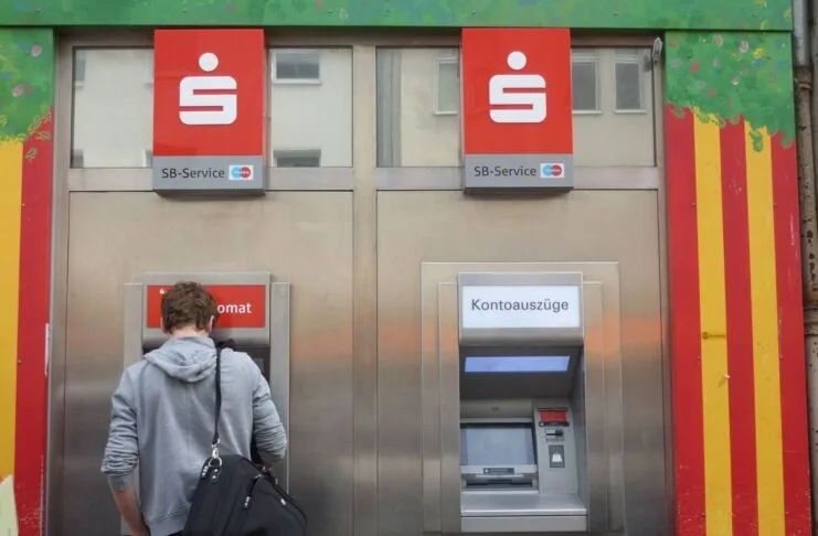 Geldautomat Sparkasse Kunde Bank Bargeld Kontoauszug Filiale Bankkarte Ein Mann mit grauem Kapuzenpullover und schwarzer Umhängetasche steht an einem Geldautomaten einer Sparkasse. Links befindet sich der Geldautomat, rechts daneben ein Kontoauszugsdrucker.