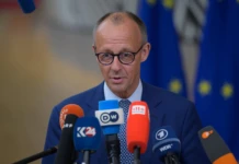 Bundeskanzler Friedrich Merz, der eine Brille, eine Krawatte und einen blauen Anzugsakko trägt, steht vor einer Reihe von Mikrofonen verschiedener Medienvertreter. Im Hintergrund sind EU-Flaggen zu sehen.