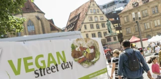 Auf einem großen Platz in einer Stadt stehen verschiedene Zelte sowie mehrere unterschiedliche Imbissbuden. Menschen bewegen sich zwischen den Ständen und den Zelten. Im Vordergrund steht ein großer Banner, auf dem "Vegan Street Day" steht.