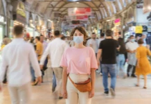 Eine junge Frau steht mit einem Mundschutz an einem öffentlichen Ort, um sich und andere vor einer Ansteckung mit Viren oder Bakterien zu schützen. Um sie herum tummeln sich viele andere Menschen.
